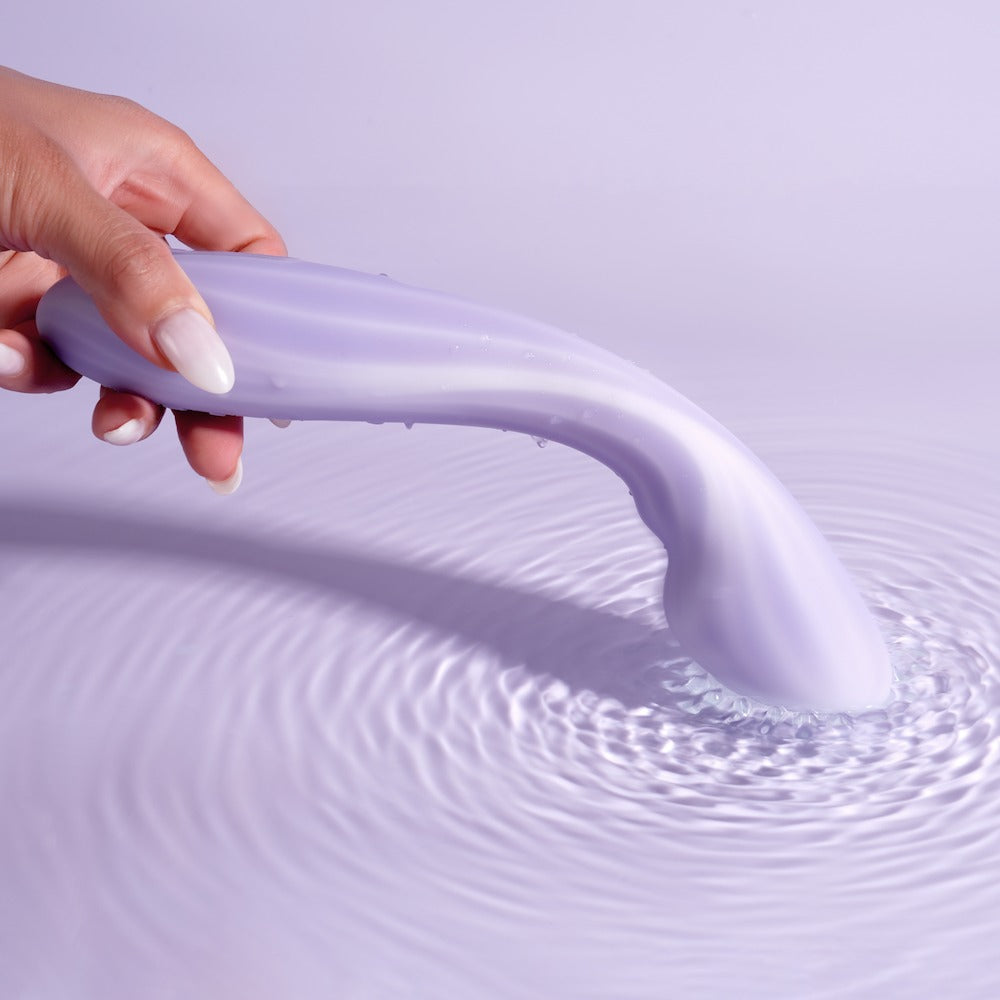 Hand holding the Svakom Margot Dual-Motor Warming G-Spot Vibrator over water with vibration ripples on a light purple background