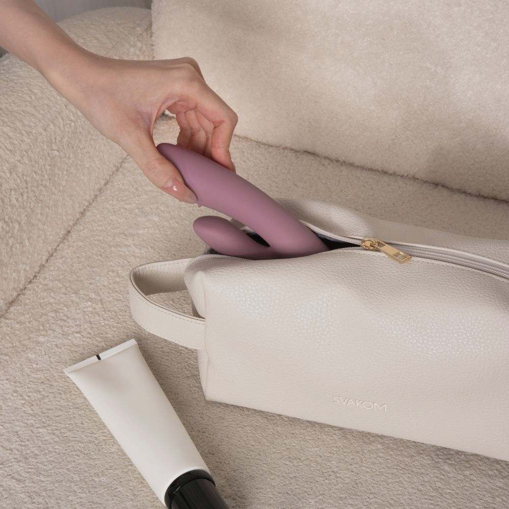 Svakom | Vegan Leather Storage Bag with Carry Handle (Beige) with a purple rabbit vibrator being placed inside, featuring the embossed 'SVAKOM' logo and gold-tone zipper on a cream textured surface