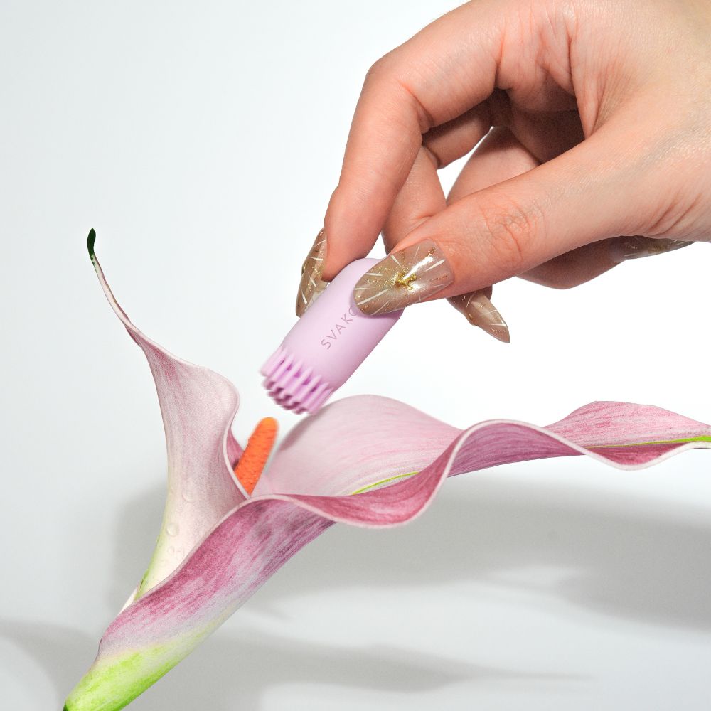 Brush-tip end of Svakom Plume Interactive Brush Tail Bullet Vibrator in pastel lilac held above a flower, showing the soft silicone brush texture and flexible tail.