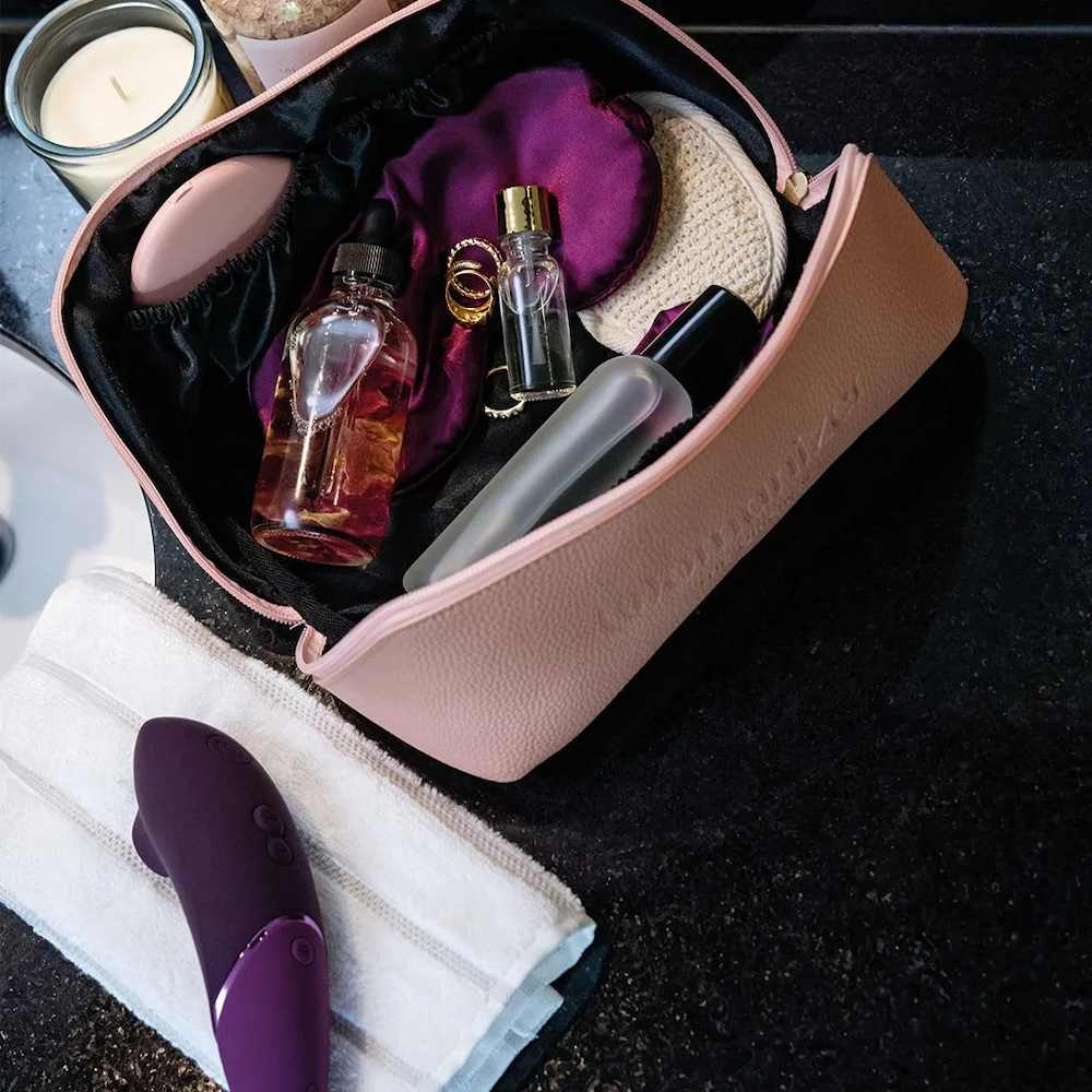 Open view of the blush pink Womanizer Cosmetic Toy Bag with Zip & Carry Handle filled with assorted intimate wellness items, including a purple Womanizer toy, perfume bottles, a cream sponge, and a purple eye mask, arranged on a dark countertop next to a folded white towel